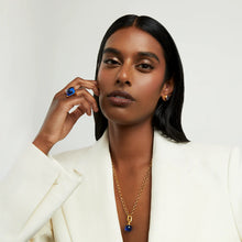 Load image into Gallery viewer, A woman with long black hair and a serious expression wears a white blazer, posing with her left hand framing her face. She showcases a gold necklace with a blue pendant and an elegant Plaza Ring adorned with hand-faceted semi-precious gemstones, all against a plain white background.
