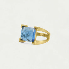 Load image into Gallery viewer, The Mini Plaza Ring features a hand-faceted rectangular blue gemstone held by four gold prongs. Its polished band splits at the shoulders, offering a modern look, while its light backdrop enhances the ring&#39;s beauty.
