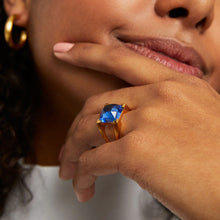 Load image into Gallery viewer, With curly hair, a person wears gold hoop earrings and a Mini Plaza Ring showcasing a bold blue gemstone on their index finger. Their chin rests on pale pink polished nails as they wear a white top in front of a softly blurred background.
