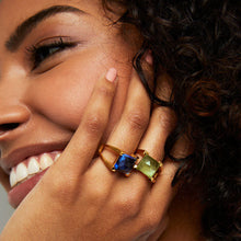 Load image into Gallery viewer, A woman with curly hair beams a smile, displaying her Mini Plaza Ring with one blue and one green semi-precious gemstone as she rests her hand on her cheek. Her light pink nails complement the jewelry and highlight her joyful expression.
