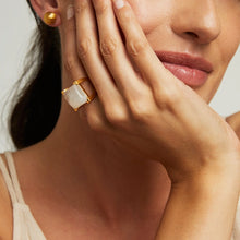 Load image into Gallery viewer, A light-skinned woman with her hair pulled back rests her chin on her hand, displaying a serene expression as she showcases the magnificent Plaza Ring on her ring finger. The ring features a hand-faceted semi-precious gemstone, paired with gold stud earrings and a beige tank top, while she wears a slight smile.
