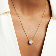 Load image into Gallery viewer, A close-up shows a person wearing a delicate gold chain necklace featuring the Signature Pendant. This iridescent man-made gemstone elegantly rests between their collarbones, framed by a cream-colored V-neck garment.
