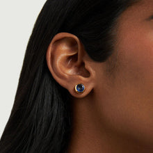 Load image into Gallery viewer, A close-up shows a person&#39;s ear adorned with Dean Davidson&#39;s Signature Midi Studs, featuring a dark blue man-made gemstone in a gold-tone bezel. The straight, dark hair partly covers the ear against a light background.
