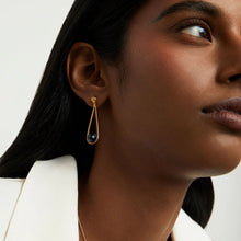 Load image into Gallery viewer, A person with long, dark hair wears the Mini Ipanema Earrings, featuring ethically sourced dark blue gemstones set in gold teardrop frames. Their white blazer and the light background enhance the close-up focus on this elegant accessory and their profile.
