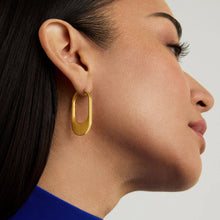 Load image into Gallery viewer, A person with long dark hair is seen in profile, wearing Crosby Hinged Hoops, crafted in brushed 18kt gold with an oval-shaped open center. Dressed in a royal blue top, the light gray backdrop showcases the earrings&#39; modern edge and sleek design.
