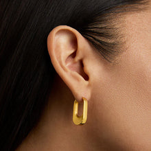 Load image into Gallery viewer, Close-up of a person&#39;s ear wearing the DD Signature Crosby Hinged Huggie Hoops in brushed 18kt gold, featuring an elongated U-shape with a polished finish. Dark hair is swept back, contrasting against warm skin tones and a neutral background that emphasizes the earring.
