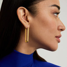 Load image into Gallery viewer, A person with long dark hair wears Crosby Drop Hoops crafted from 18kt gold. Dressed in a vibrant blue turtleneck, their profile and earrings are elegantly highlighted against a neutral light gray background.
