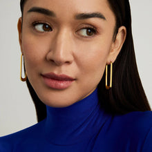 Load image into Gallery viewer, Wearing a royal blue turtleneck, a person with long dark hair looks to the side, subtly expressing themselves. They wear minimalist Crosby Drop Hoops in 18kt gold, hanging elegantly alongside their face against a plain background that highlights their refined features.
