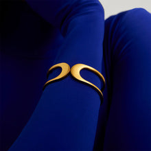 Load image into Gallery viewer, A close-up of the Crosby Midi Cuff on a person&#39;s arm, showcasing its crescent-shaped design with a 22-Karat Gold Finish. The lightweight cuff beautifully contrasts against form-fitting royal blue fabric, creating a striking visual effect.
