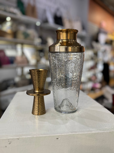 The Stylish Bar Gift Set features a textured glass cocktail shaker with a hammered bronze lid and a matching double jigger on a white surface, with blurred shelves of bar essentials in the store backdrop.