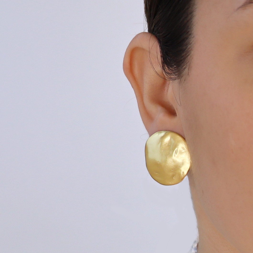 Close-up of a person wearing large, circular Shell Disc Clip-On Earrings with a hammered texture. These 24-kt gold-plated earrings feature subtle indentations and are highly reflective. The person's ear and part of their cheek are visible against a neutral background.