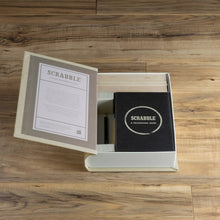 Load image into Gallery viewer, An open box on a wooden floor showcases a Scrabble Vintage Bookshelf Edition, featuring an elegantly designed lid with Scrabble-themed text. Inside, the premium game components are neatly arranged beneath a black cover labeled &quot;SCRABBLE: A Crossword Game.
