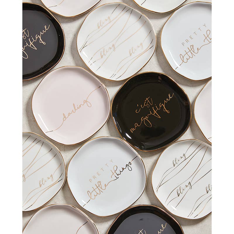 The "Trinket Tray - Little Things" collection includes small square ceramic trays with rounded edges and metallic gold rims. Featuring cursive text like "darling," "pretty little things," and "c'est magnifique," the trays come in black, white, and pale pink with an artful overlapping pattern.