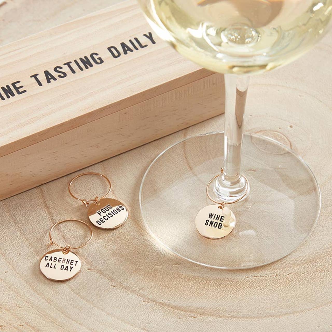 The "Wine Tasting Daily" pine box with gold wine charms, including "Wine Snob," "Poor Decisions," and "Cabernet All Day," is perfect for gifting. It sits on a light wooden surface, creating a casual and inviting wine theme.