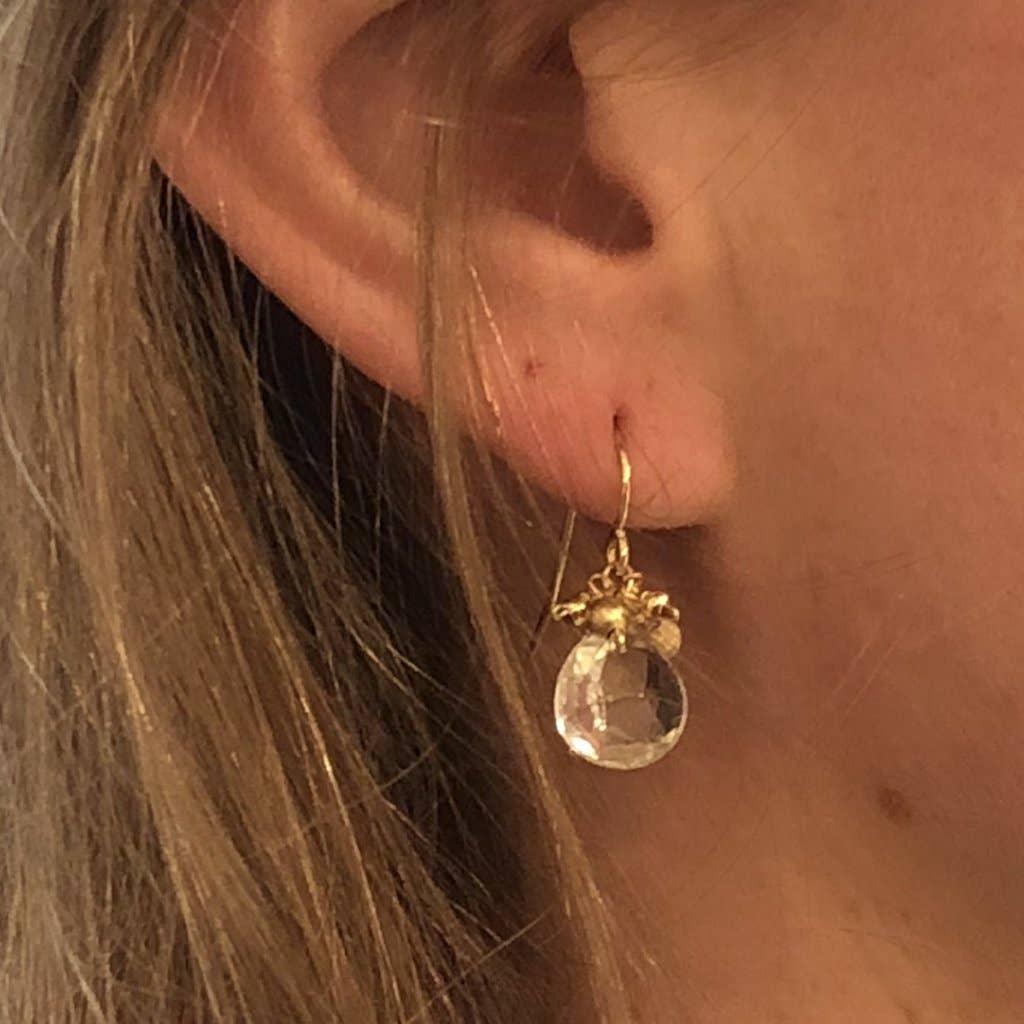 A close-up shot showcases an ear adorned with a Mini Cluster Earring: Gold-Filled, featuring a large, faceted clear stone and sapphire roundels. This drop-style earring elegantly dangles from a delicate gold hook. The lighting enhances the reflective facets as light brown hair beautifully frames the scene.