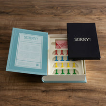 Load image into Gallery viewer, The Sorry! Vintage Bookshelf Edition is displayed, featuring an open box that reveals a vintage game board and colorful plastic pieces neatly organized. A fabric-covered book containing the rulebook and cards rests alongside the turquoise cover, which includes a sleek black interior compartment.
