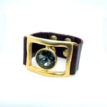 Load image into Gallery viewer, The Open Rectangle w Single Stone Leather Bracelet in Gold features a wide, dark leather band with an 18K gold-finished frame and a faceted dark blue gemstone. Handcrafted in Brooklyn, it has button studs for closure and a minimalist design against a white background.
