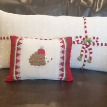 Load image into Gallery viewer, Two decorative pillows adorn a brown leather couch: a smaller one featuring an embroidered hedgehog in a red and gray hat amid festive patterns, and the Gift Lumbar Pillow, Ecru displaying a candy cane-striped bow and holly. Both exhibit the charm of handmade Armenian crafts.
