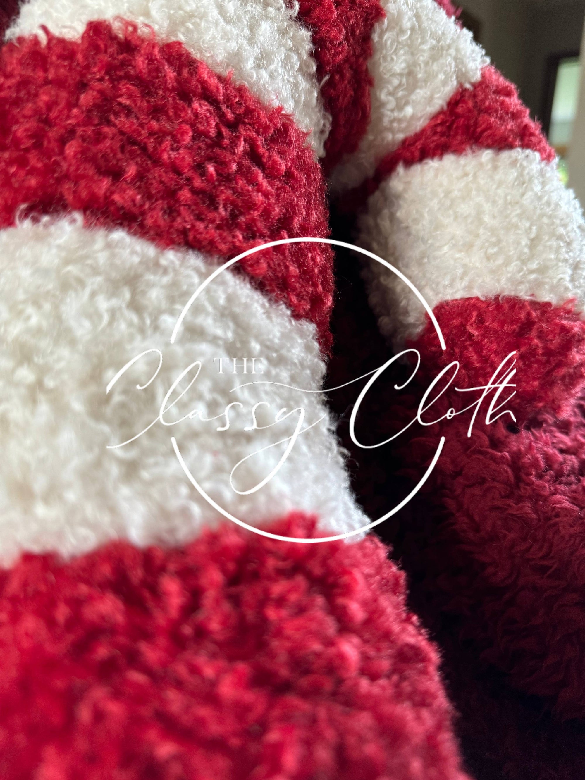 Close-up of a plush, red-and-white striped fabric that brings to mind the Red Candy Cane design of the Christmas Sherpa Pillow. The image features a circular logo with elegant script reading "The Classy Cloth." The texture looks soft and cozy, ideal for providing warmth and comfort as part of holiday decorations.