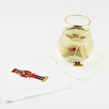 Load image into Gallery viewer, A glass with a gold rim holds a light-colored beverage on a napkin from the Nutcracker Embroidered Cocktail Napkin Boxed Set. Next to it, another 100% cotton napkin showcases the vivid red and gold nutcracker design. The elegant plain white surface in the background accentuates the festive charm.
