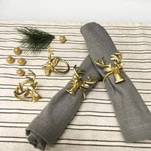 Load image into Gallery viewer, Two gray napkins, crafted by hand from sustainable materials, are rolled with Gold Deer Napkin Rings (Set of 4) on a striped cloth background. Additional gold deer rings, small golden ornaments, and a sprig of pine nearby complete the festive holiday display.
