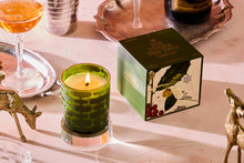 Load image into Gallery viewer, A decorative holder embraces the 5.5 oz &quot;Wine Under The Tree&quot; candle, its green glow illuminating a table adorned with its accompanying box. Nearby, a silver tray, a gold deer figurine, and a cocktail glass filled with an amber liquid create an elegant display. The subtle cedar aroma enhances the festive ambiance.
