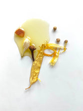 Load image into Gallery viewer, A glossy puddle of honey glistens on a white surface, topped with a garlic clove, a piece of dried orange, a clove, and a golden leaf surrounded by scattered small seeds. This natural arrangement evokes the therapeutic aroma reminiscent of &quot;Need Sleep&quot; - Aromatherapy Body Oil.
