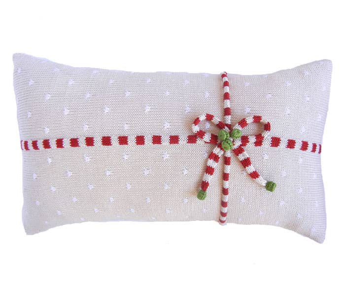 The Gift Lumbar Pillow, Ecru, is a handmade rectangular creation showcasing white polka dots and embellished with a red and white knitted striped ribbon tied into a bow, accentuated with small green leaves. Its design radiates a festive holiday charm inspired by traditional Armenian crafts.