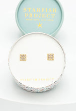 Load image into Gallery viewer, A pair of hypoallergenic Dignity Zircon Stud Earrings, crafted in 14K gold, is nestled in a decorative round box. The lid, tilted behind the box, is light blue with &quot;Starfish Project&quot; written in gold letters alongside a small starfish icon. The inner lining of the box is white.
