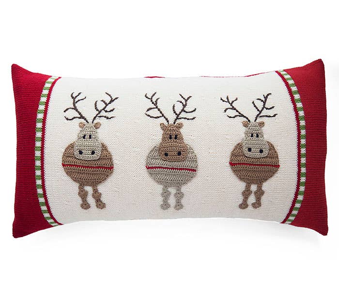 This festive Round Reindeer Lumbar Pillow features a vibrant red border and displays three knitted reindeer set against a cream background. Each reindeer is designed with a round body, antlers, and dangling legs. The edges of the pillow are decorated with stripes in green, red, and cream hues. This piece beautifully exemplifies handmade craftsmanship with Armenian influences.