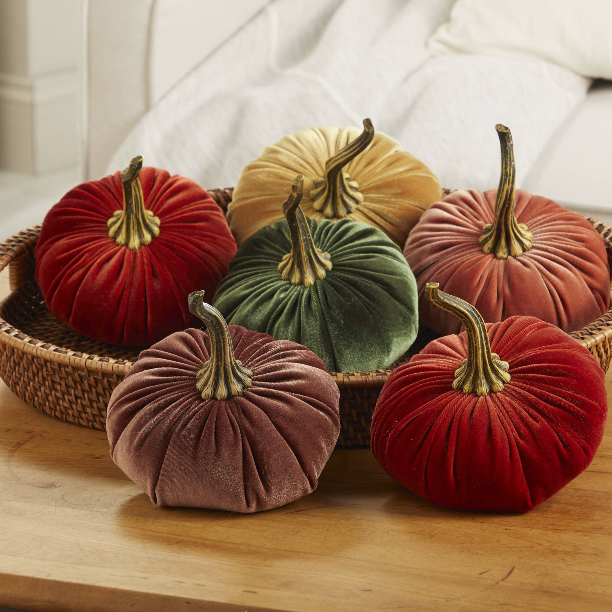A woven basket on a wooden table holds six Handmade Large Velvet Pumpkins. The rich hues of red, orange, brown, gold, and green showcase realistic brown stems. Set against the backdrop of a white sofa, the arrangement perfectly captures the essence of cozy fall decorating at its best.