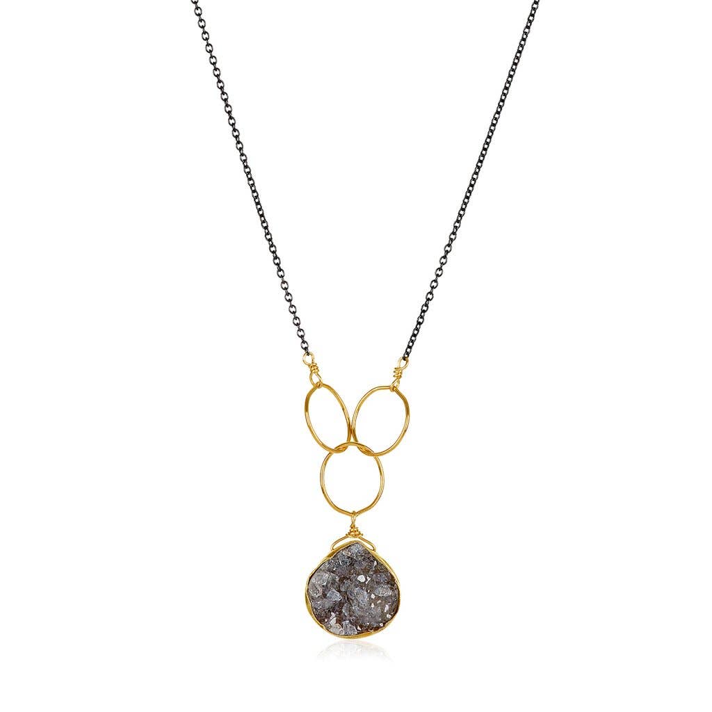 The Cassiopeia Necklace boasts a black gold-filled chain and features a pendant of interlinked gold loops, which elegantly hold a textured, irregularly shaped stone. This modern design beautifully showcases the stone in its gold setting at the base of the loops and is prominently displayed against a white background.