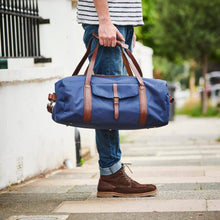 Load image into Gallery viewer, A person wearing blue jeans and brown boots stands on a sidewalk, holding a Leather Trim Weekend Travel Bag with brown leather straps. The background features a blurred street scene with a white wall and greenery, accentuating the bag and lower attire while keeping the upper body out of view.
