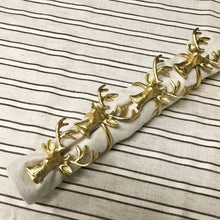 Load image into Gallery viewer, A set of four gold deer napkin rings, each exhibiting intricate antlers and lifelike textures, holds a white napkin adorned with brown stripes. These handmade rings are beautifully displayed in a row against a sustainable fabric backdrop featuring alternating black and white lines.
