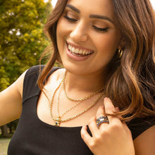 Load image into Gallery viewer, A smiling woman with long, wavy brown hair wears a black top and layered gold necklaces. With one hand, she holds her hair back, showcasing the Gold Plated Spaced Baguette Eternity Ring on her finger. The background is blurred but reveals lush green trees and grass, suggesting an outdoor setting.
