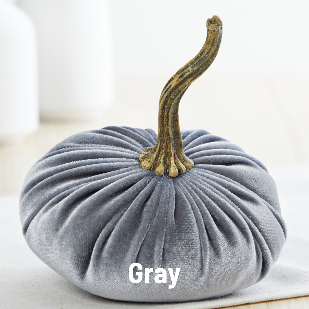 Displayed on a light-colored wooden surface is a plush, handmade small velvet pumpkin in gray, featuring a textured, wrinkled surface and a natural brown stem. In the background, two white cylindrical objects are partially visible out-of-focus. This piece is perfect for fall decorating or as an elegant wedding centerpiece.