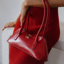 Load image into Gallery viewer, A person in a red dress holds a structured, rectangular Dakota Recycled Vegan Shoulder Bag with gold hardware. This shiny handbag rests near their hip against a soft, neutral background that contrasts beautifully with the vibrant ensemble and its OEKO-TEX certified lining.
