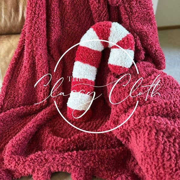 A plush Christmas Sherpa Pillow featuring a Red Candy Cane design is cozily placed on a brown leather chair, adding charm to the ambiance. The words "The Classy Cloth" are gracefully overlaid in white script within a circular design.