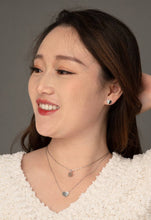 Load image into Gallery viewer, A woman with long brown hair smiles while looking to the right. She is wearing heart-shaped earrings and two delicate necklaces, including the Alexis Silver Heart Necklace with a small circular pendant. Her white textured top complements her subtle makeup against a gray background.
