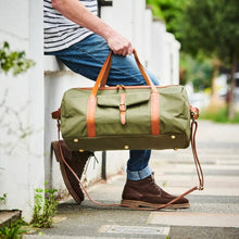 Load image into Gallery viewer, A person in blue jeans and brown boots sits on a white wall, holding the Leather Trim Weekend Travel Bag. This water-resistant green canvas bag has brown leather straps, gold hardware, and features a front flap pocket. They are outdoors on a pavement with greenery and trees in the background.
