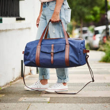 Load image into Gallery viewer, A person in cuffed jeans and white sneakers stands on a sidewalk holding the Leather Trim Weekend Travel Bag, a large blue travel bag with brown leather straps and a front pocket. This water-resistant bag is equipped with an attached shoulder strap, while the background shows a blurred residential street and a white wall.
