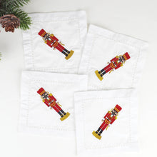 Load image into Gallery viewer, A boxed set of Nutcracker Embroidered Cocktail Napkins showcases four 100% cotton napkins, each adorned with a red and gold nutcracker design. A sprig of pine with a small pinecone in the top left corner enhances their festive appeal. These machine washable napkins seamlessly combine practicality with elegance.
