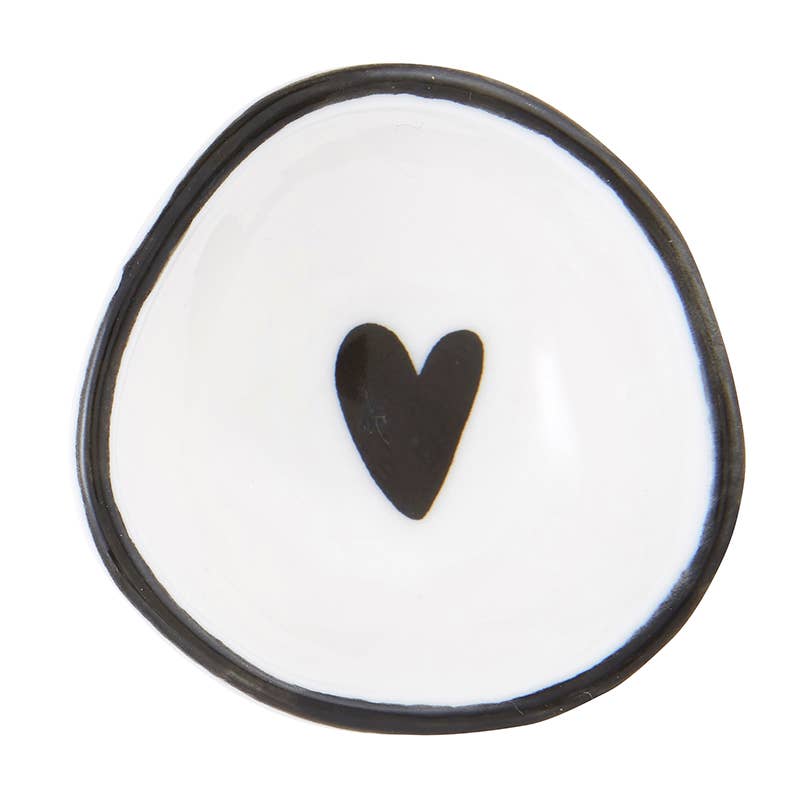 The Ring Dish is a small, white ceramic piece with a slightly irregular round shape, featuring a black heart in the center and a black rim. Its glossy finish adds to its elegant handmade look, perfect for storing rings.