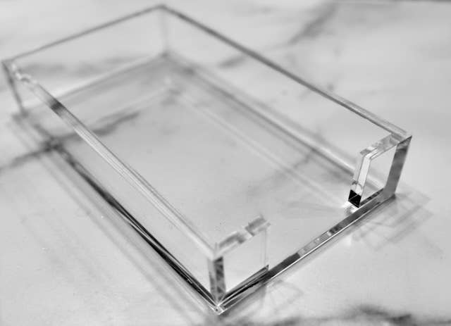 Acrylic Guest Towel Tray – a clear, rectangular piece with open top and sides, gracefully perched on a marble surface. This modern accessory showcases smooth, transparent edges and corners that create delicate shadows and reflections. Its softly blurred background highlights the tray's simple elegance and sophistication.