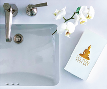 Load image into Gallery viewer, On the left is a bathroom sink with a silver faucet. To the right, a white book with gold text and an illustration of a meditating figure reads &quot;LET THAT SHIT GO.&quot; Above neatly folded Cloth-Like Guest Towels - Year Round Themes, a white orchid branch adds elegance to the smooth background.
