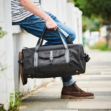 Load image into Gallery viewer, A person in blue jeans and a striped shirt sits on a low white wall, holding a large Leather Trim Weekend Travel Bag. Their feet, wearing brown shoes, rest on the sidewalk. The scene is outdoors, surrounded by greenery and a blurred background.
