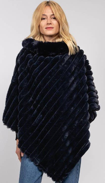 A woman with blonde hair is modeling the Faux Fur Poncho, a dark blue, plush piece made from faux fur. It features a ribbed diagonal pattern and a high, soft collar. She is posed against a plain gray background with her arms resting at her sides, showcasing stylish warmth while wearing blue jeans underneath the poncho.
