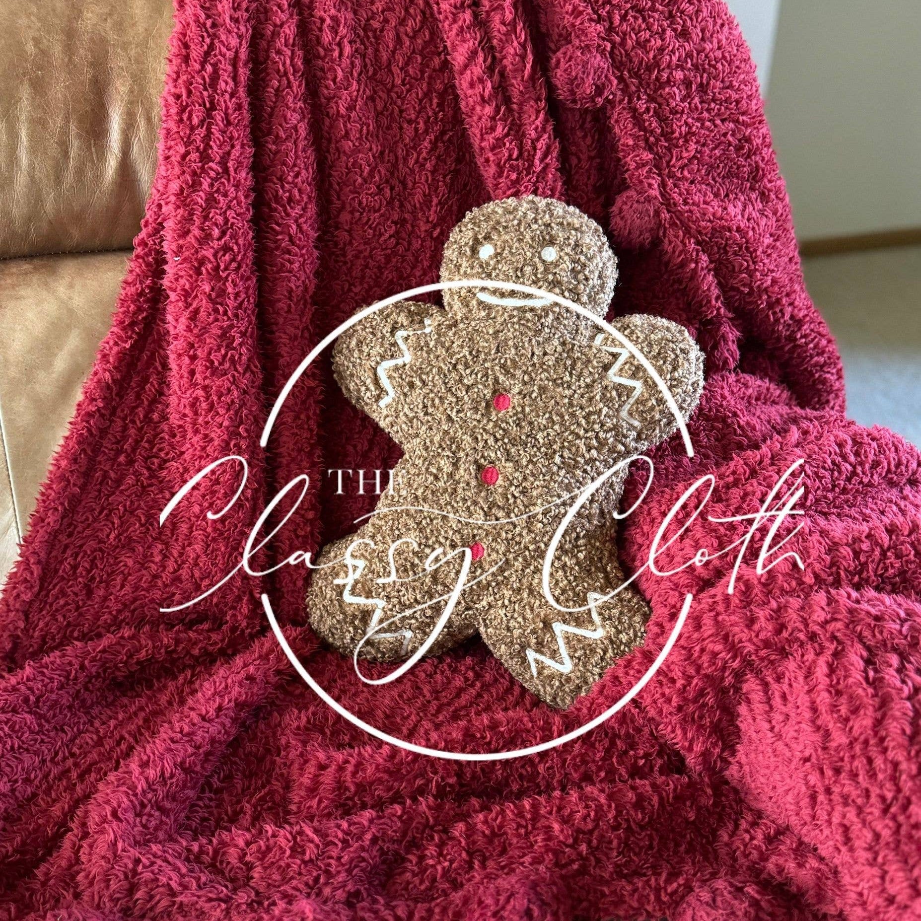 A cozy, textured red blanket is elegantly draped over a brown leather armchair, showcasing pom-pom detailing along the edges. Resting on it is a Christmas Sherpa Pillow Gingerbread Man, decorated with red buttons and white icing. The overlay text in a circular design reads "The Classy Cloth.