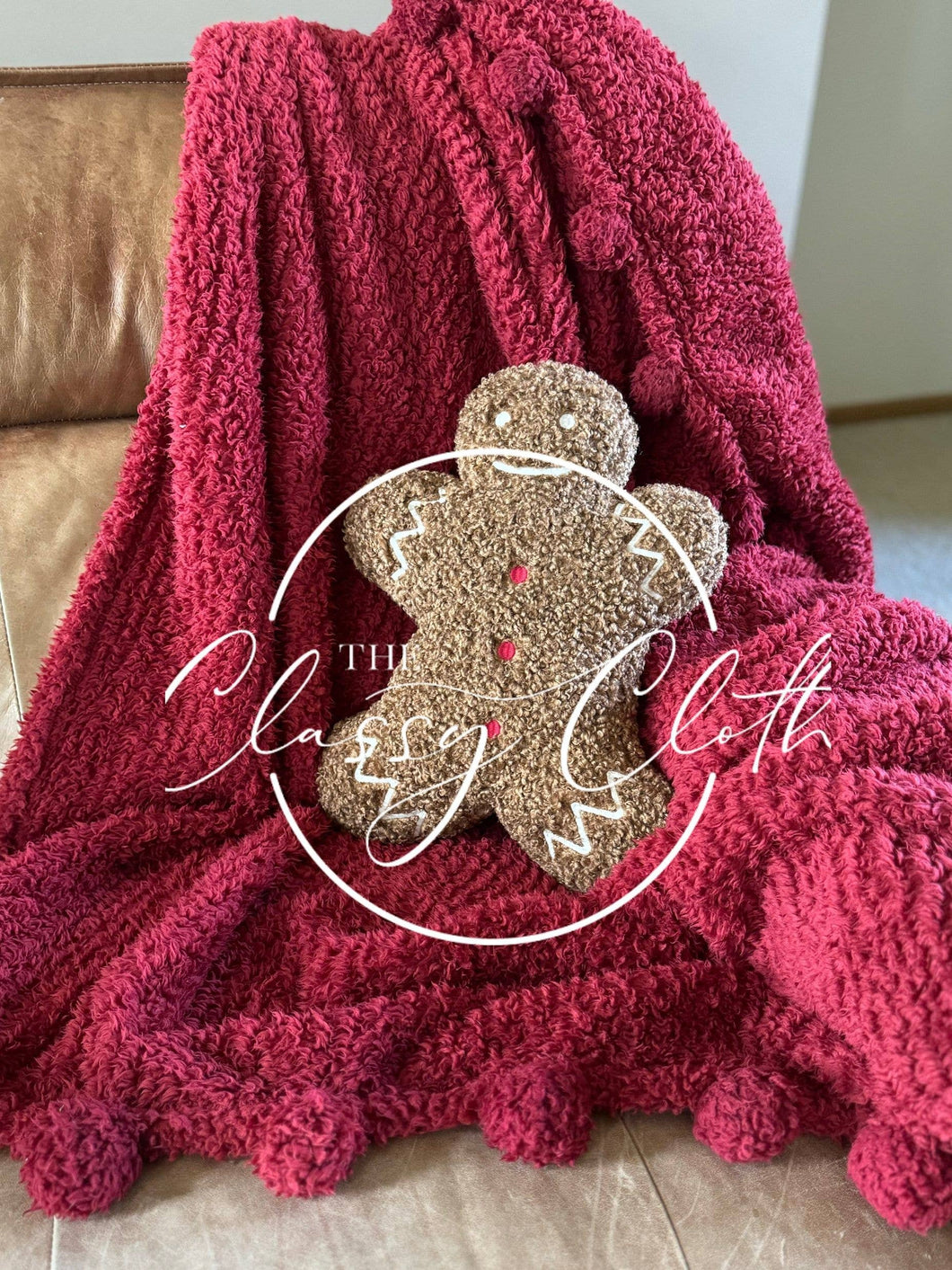 A cozy, textured red blanket is elegantly draped over a brown leather armchair, showcasing pom-pom detailing along the edges. Resting on it is a Christmas Sherpa Pillow Gingerbread Man, decorated with red buttons and white icing. The overlay text in a circular design reads 