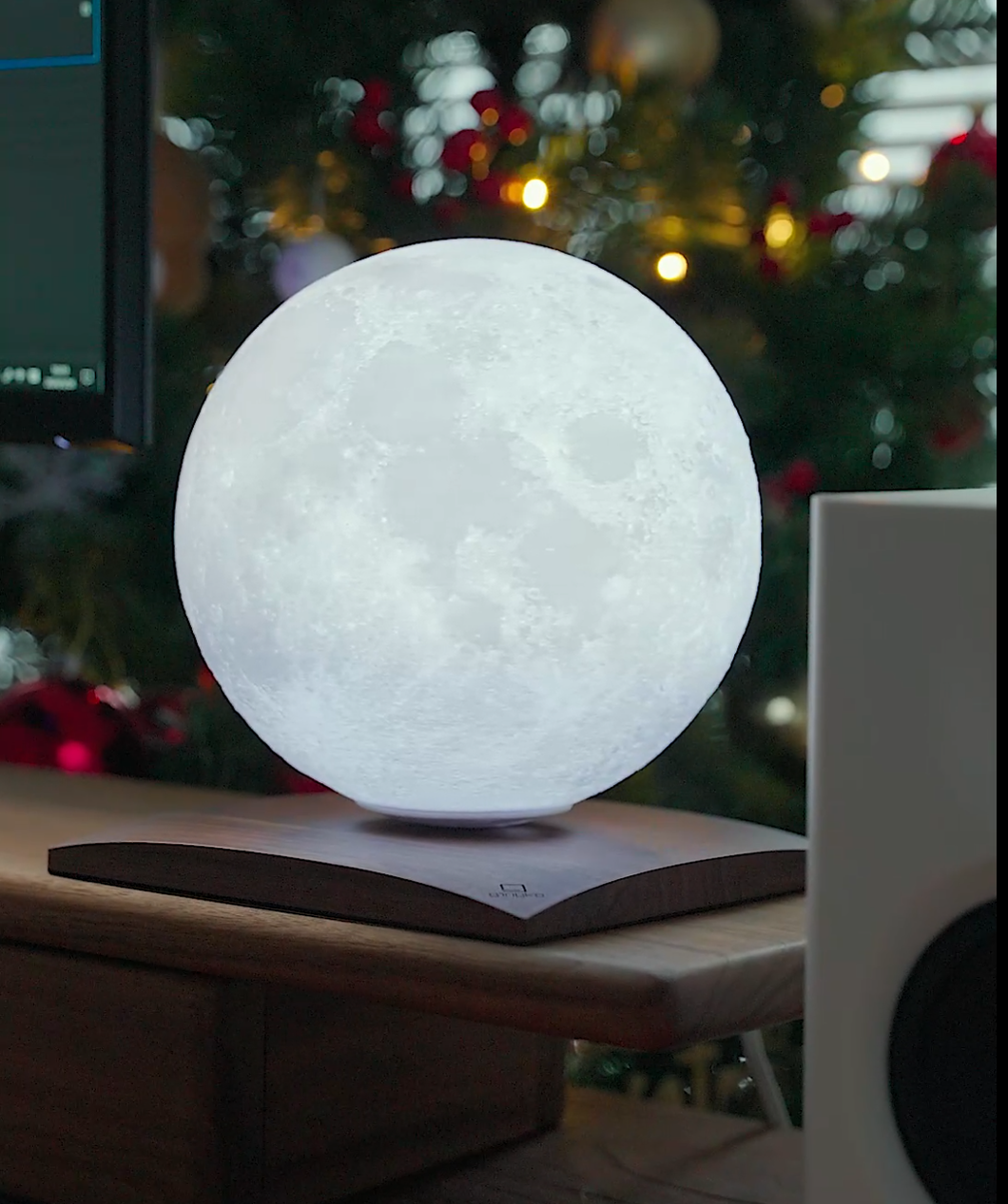 A radiant Smart LunaSpin Lamp levitates over a wooden base on a desk, casting light against a softly focused backdrop of a Christmas tree adorned with red and gold ornaments. To the right, part of a white speaker appears, enhancing the scene's cozy atmosphere with its magnetic wireless charging feature.